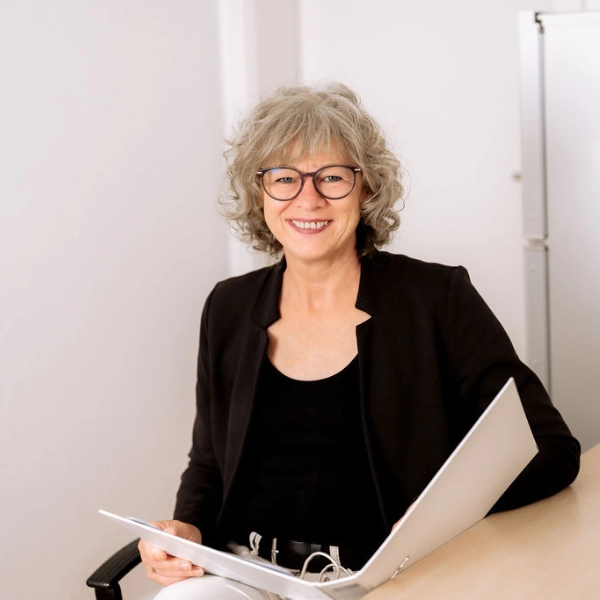 Eine Frau mit hellen, schulterlangen Haaren und einer dunklen Brille sitzt an einem Schreibtisch, lächelnd mit einem weißen Aktenordner geöffnet auf dem Schoß.